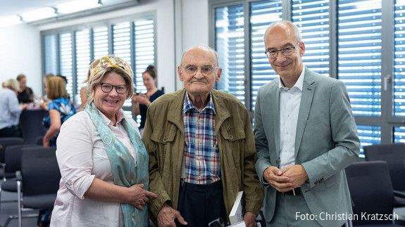 Drei Menschen stehen in einem Tagungsraum und blicken in die Kamera