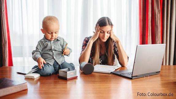 Erwerbsarbeit und private Care-Arbeit müssen zusammen gedacht werden Eine sichtlich gestresste Mutter versucht zu Hause am Laptop zu arbeiten, während neben ihr auf dem Schreibtisch ein Kleinkind sitzt und mit Büroutensilien spielt.