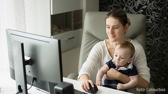 Zu sehen ist eine junge Mutter, die am Schreibtisch Homeoffice macht und ihren kleinen Sohn auf dem Arm hält.