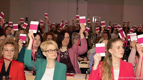 Frauen sitzen in einem großen Konferenzraum und halten Abstimmungskarten in die Luft