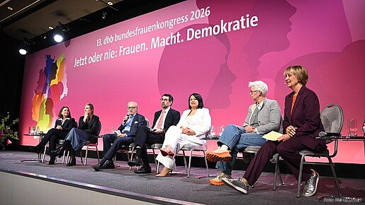 Menschen sitzen auf Stühlen auf einer Bühne und diskutieren miteinander. 