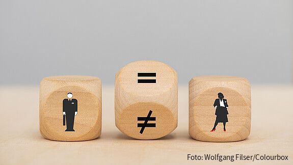 Zu sehen sind drei Würfel: Einer mit einer männlichen Figur, einer mit einer weiblichen. Der Würfel in der Mitte steht gekippt, ein Gleichheitszeichen und ein Ungleichheitszeichen sind zu sehen.