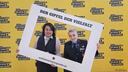 Zwei Frauen halten einen großen weißen Bilderrahmen vor einer gelben Wand