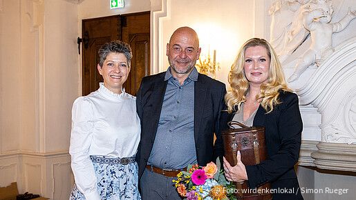 zwei Frauen und ein Mann stehen in einem Konferenzraum und blicken lächelnd in die Kamera